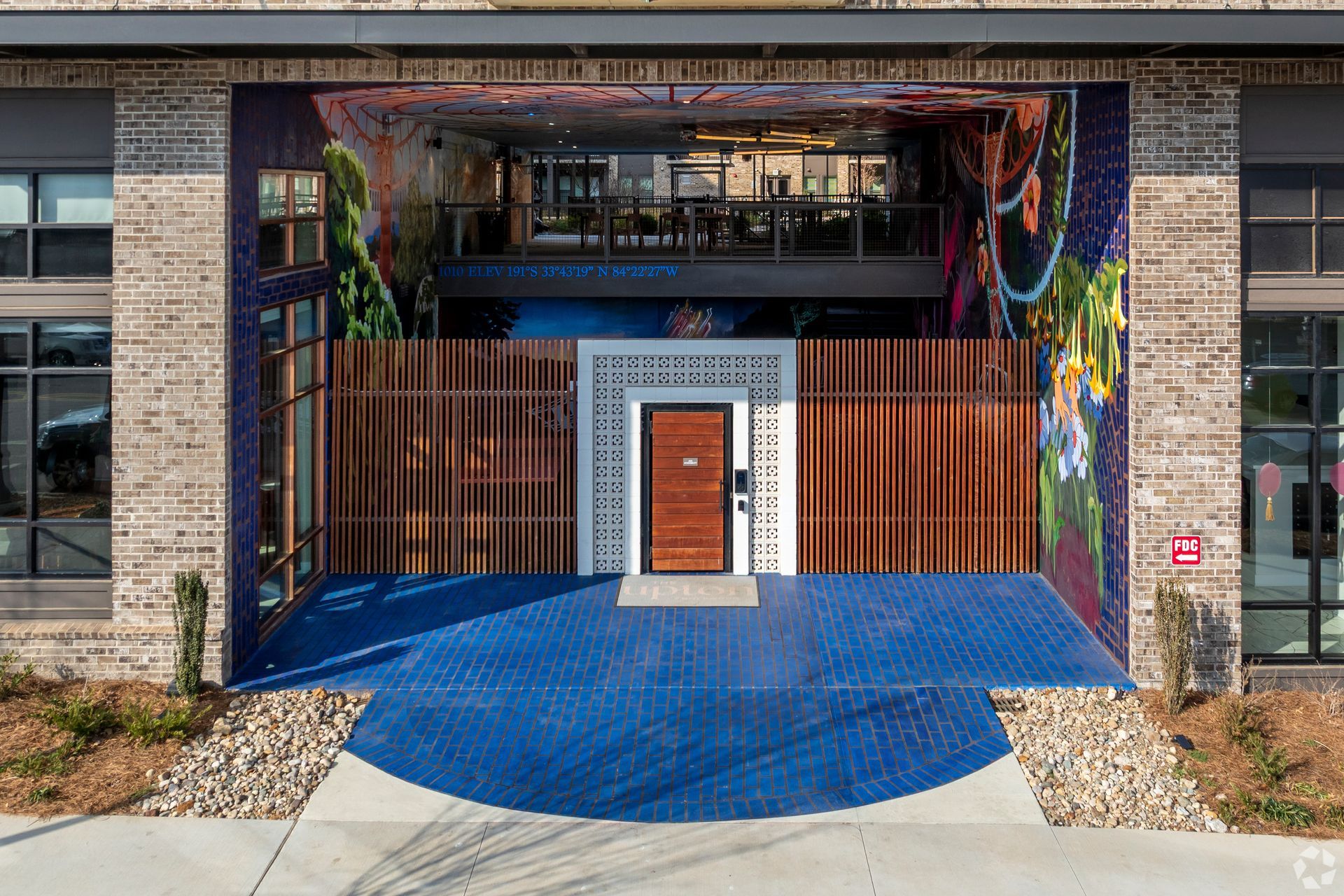 A modern brick building entrance features a central wooden door framed by patterned tile, flanked by colorful wall murals.