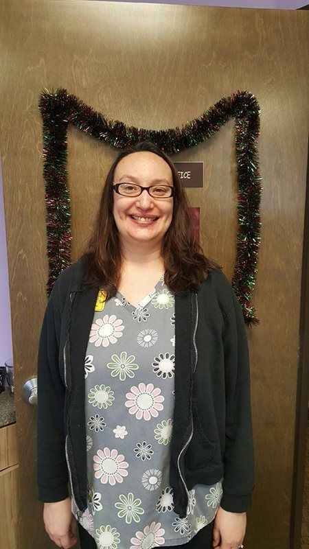 A person wearing glasses and a floral print top with a dark zip-up sweater, standing in front of a door with tinsel trim.