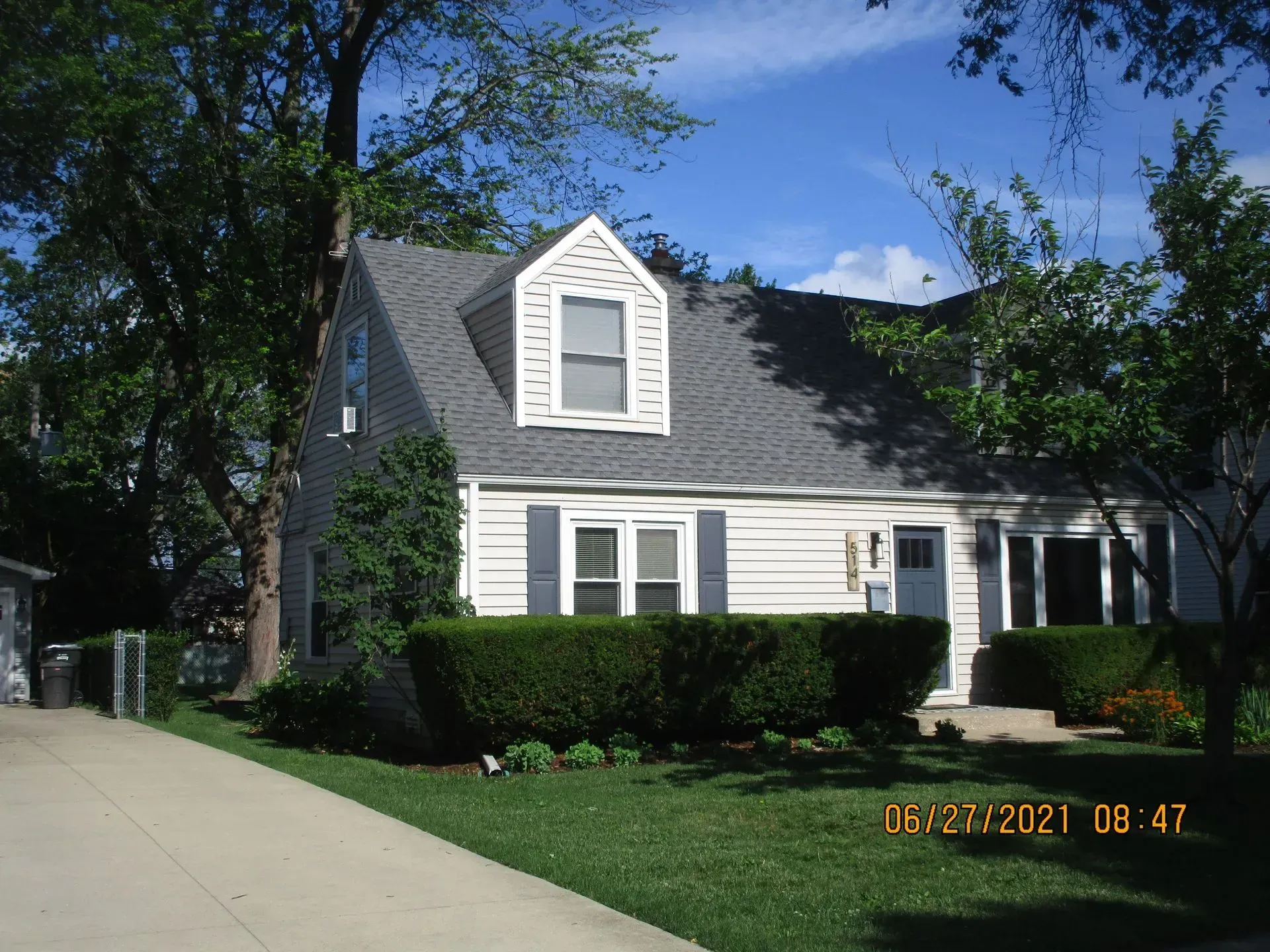 A white, single-story house with gray shingles, a dormer window, shutters, and a front lawn on a sunny day.