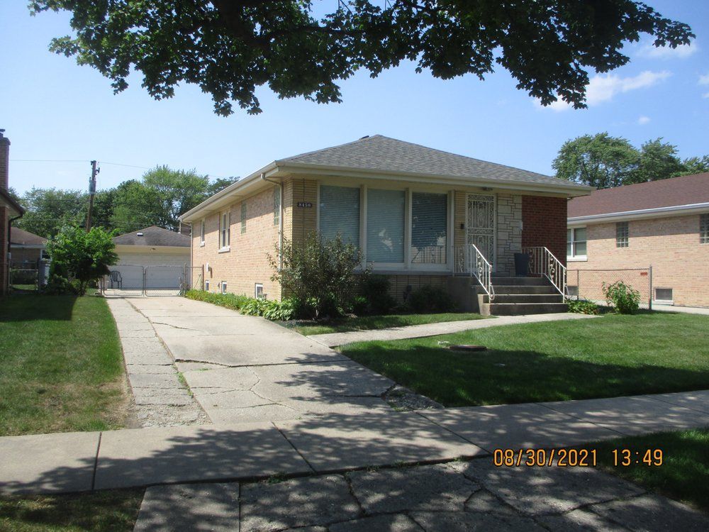 A single-story tan brick house with a front porch, concrete driveway, and a lawn under a sunny sky.