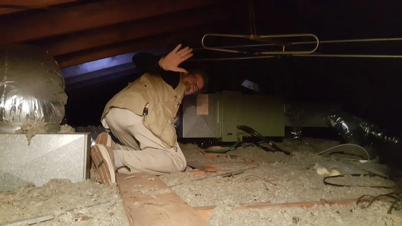 A person wearing a tan vest kneels in a dark, cramped attic near HVAC equipment and waves toward the camera.