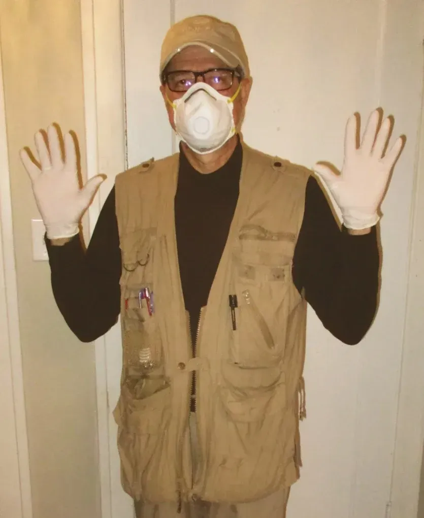 A person wearing a tan utility vest, a cap, safety glasses, a face mask, and white gloves with their hands raised.