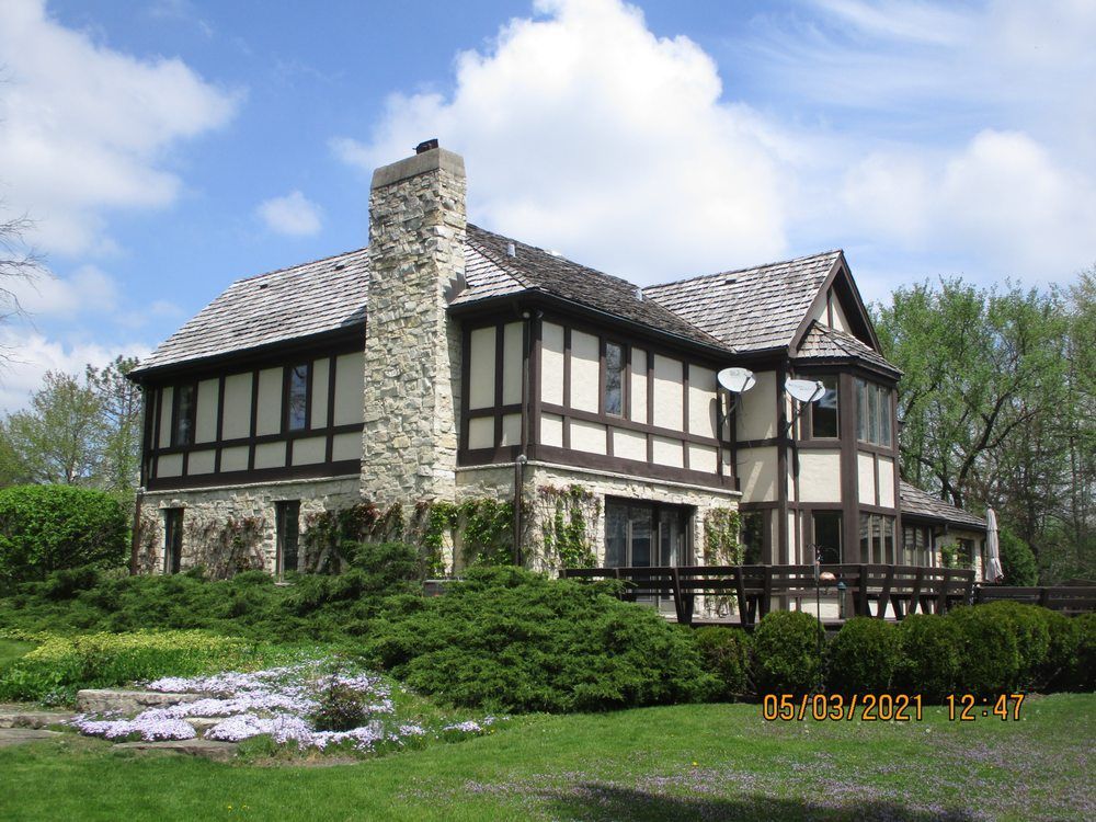 A two-story Tudor-style home featuring stone masonry, half-timbering, a prominent stone chimney, and a wooden deck.