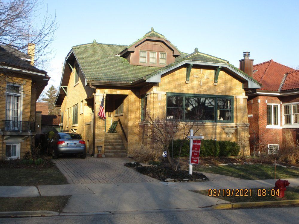 A yellow bungalow-style house with a green roof, front porch, and a car parked in the driveway on a sunny day.