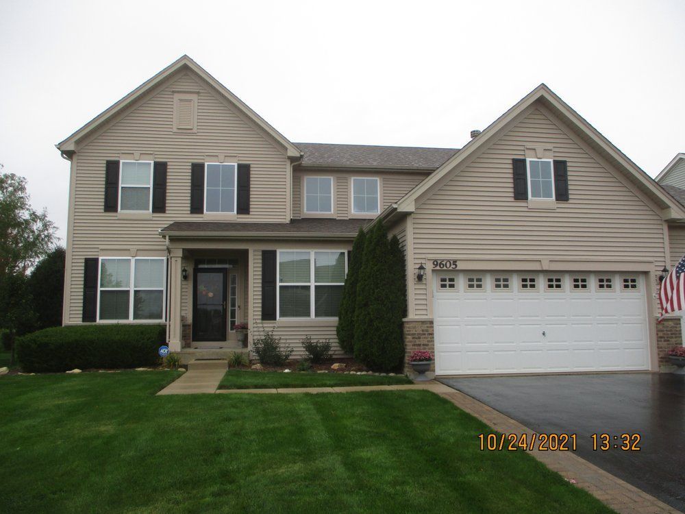 A beige two-story suburban house with a two-car garage, black shutters, and a well-maintained green lawn.