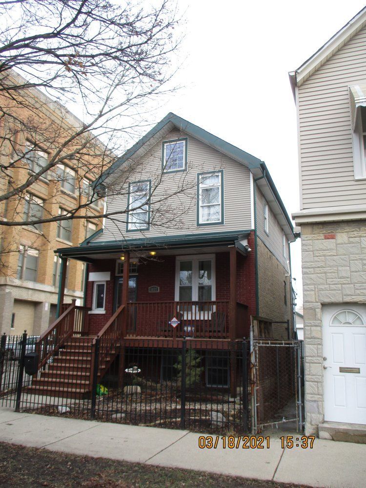 A two-story house with a wooden front porch and steps, situated between two other buildings along a concrete sidewalk.