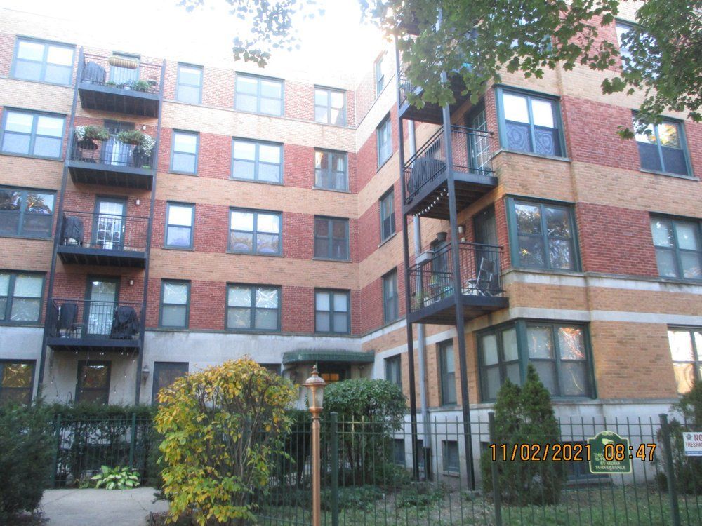 A four-story apartment building with red and tan brick, black balconies, and a metal fence in front on an autumn day.