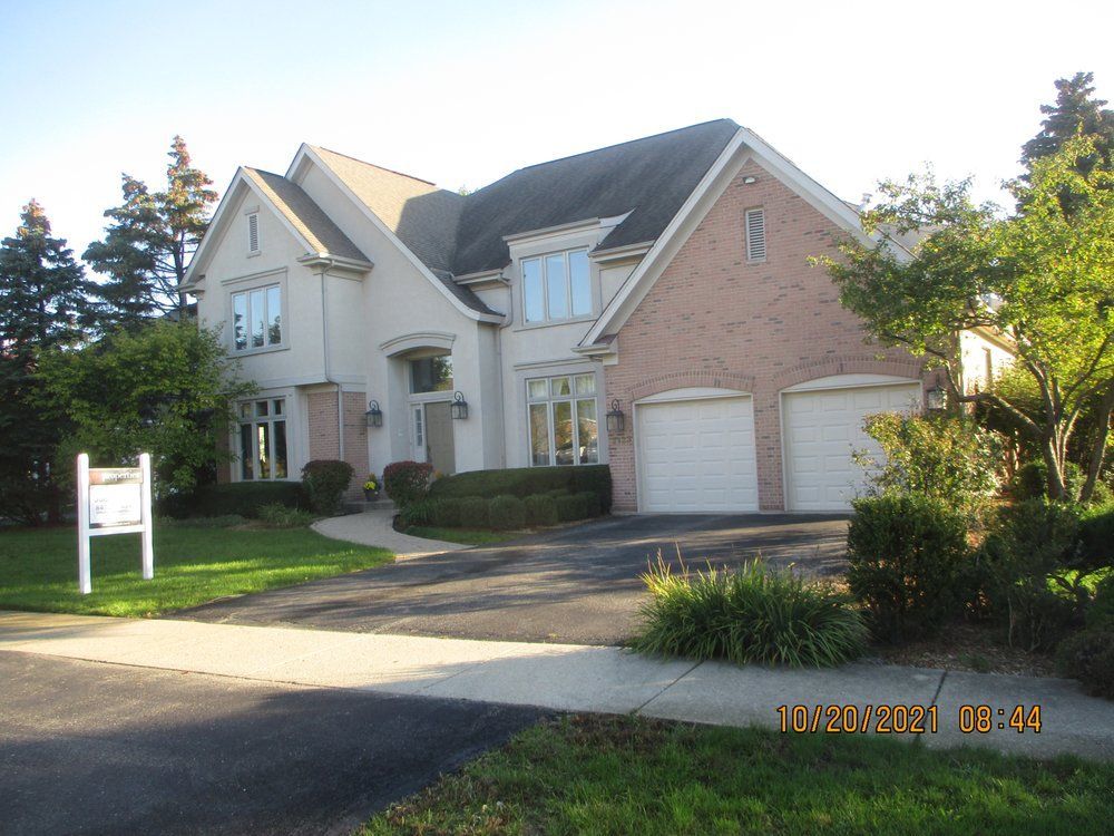 A two-story suburban house with a brick and stucco facade, a two-car garage, and a real estate sign on the lawn.