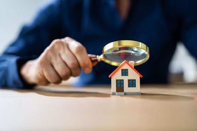 A person in a blue shirt holds a magnifying glass over a small model house on a wooden table.