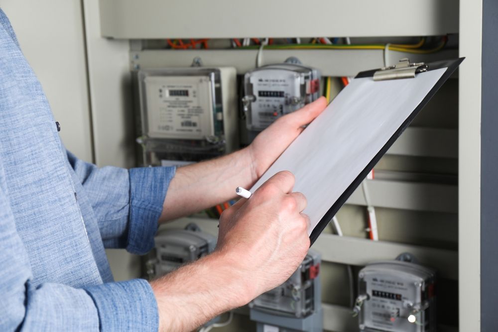 A person in a blue shirt writes on a clipboard while inspecting electrical meters in a utility box.