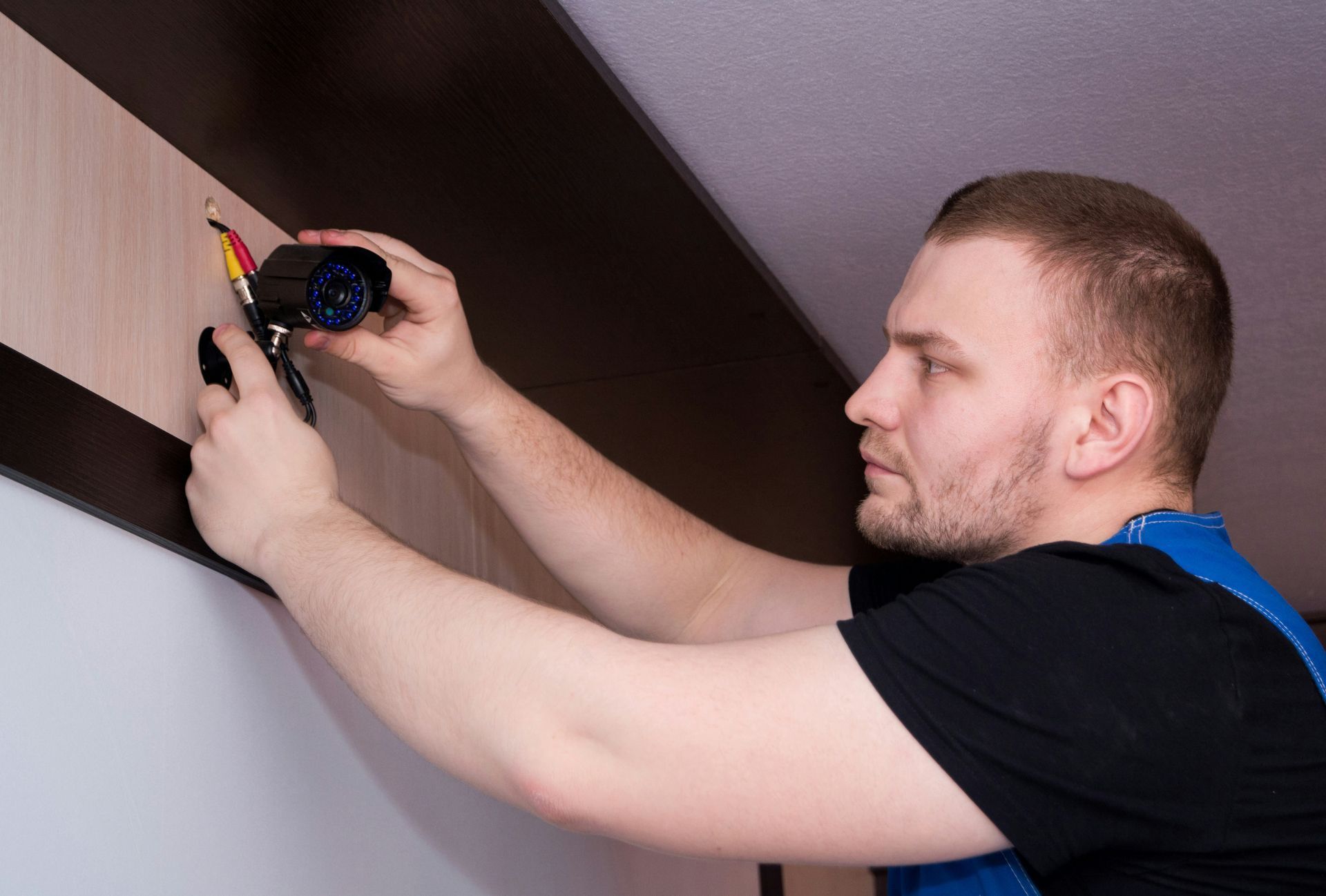 A person in a black shirt and blue overalls installs a security camera on a wood-paneled wall.