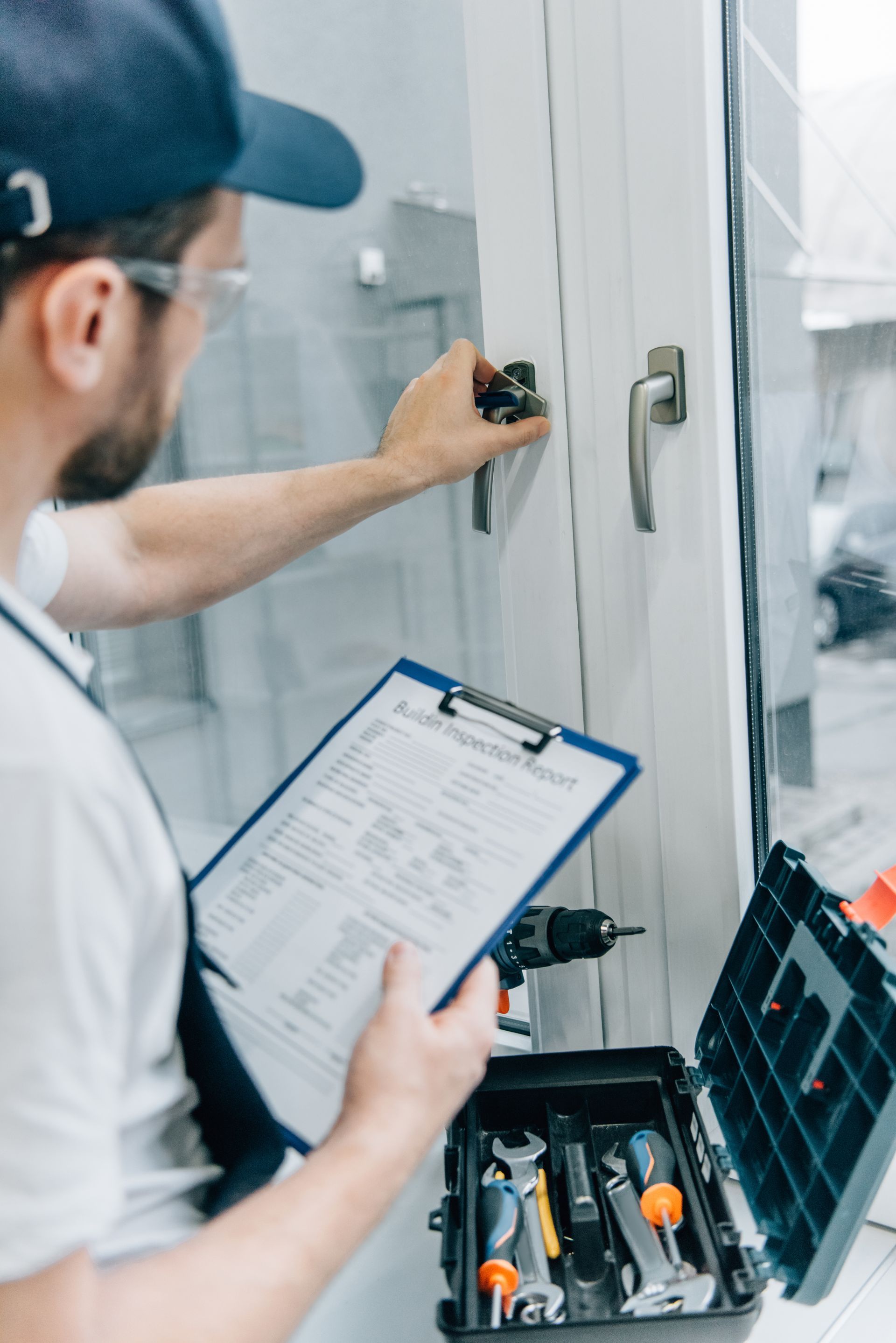 A worker in a cap and uniform examines a window handle while holding a clipboard, with a toolbox open nearby.