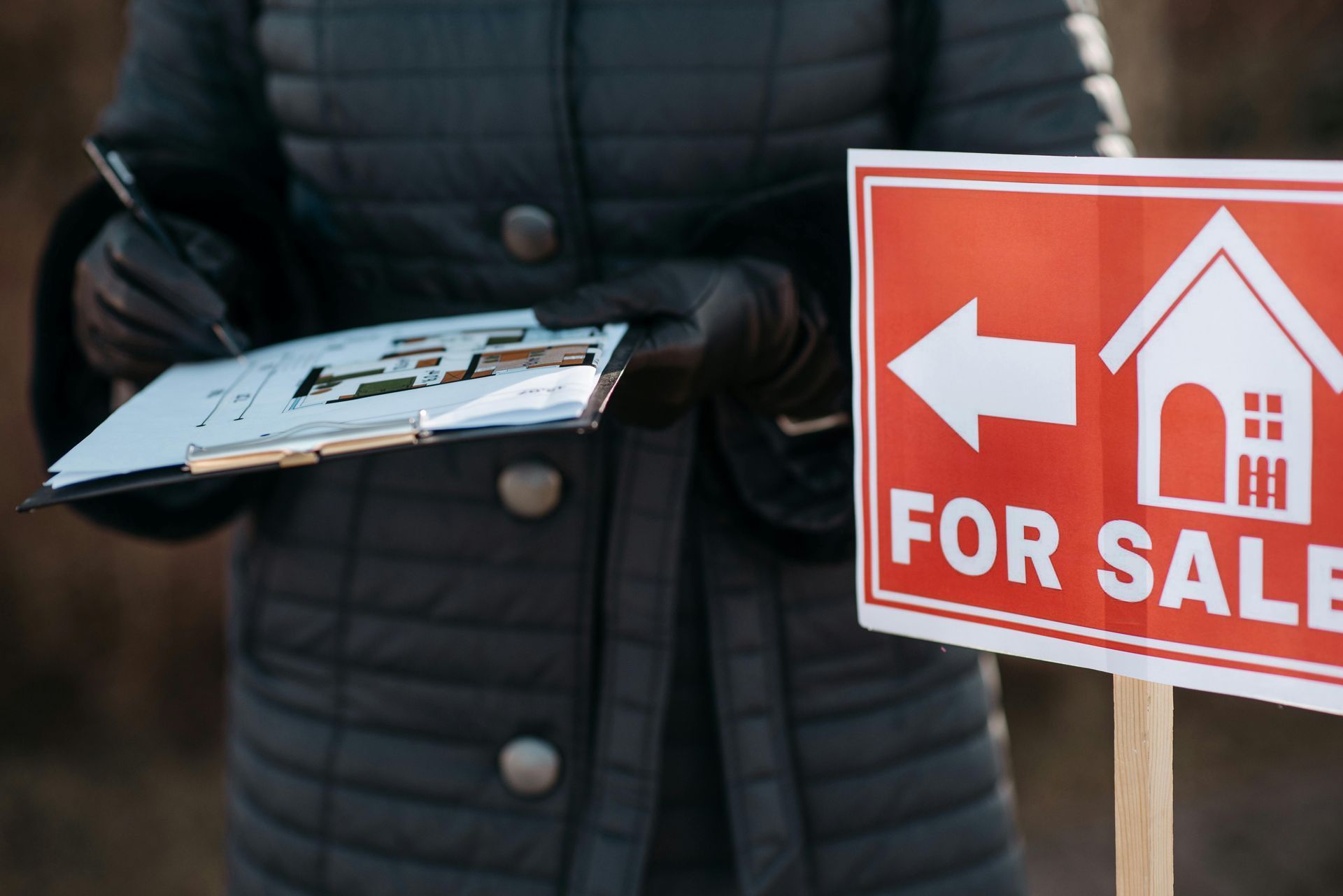A person in a black quilted coat and gloves holds a clipboard with a floor plan next to a red