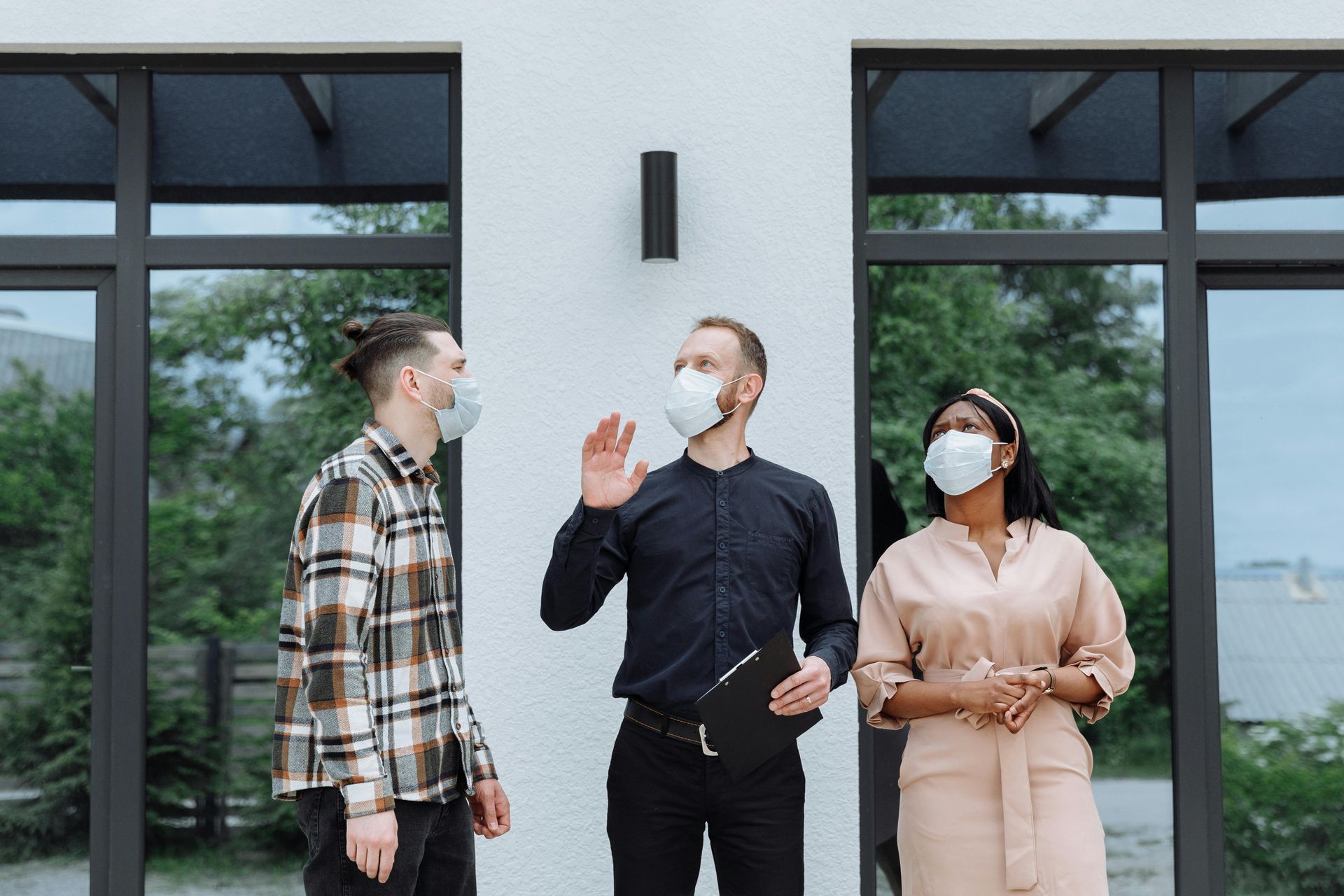 Three people wearing face masks stand outside a building; one points toward the exterior while holding a clipboard.