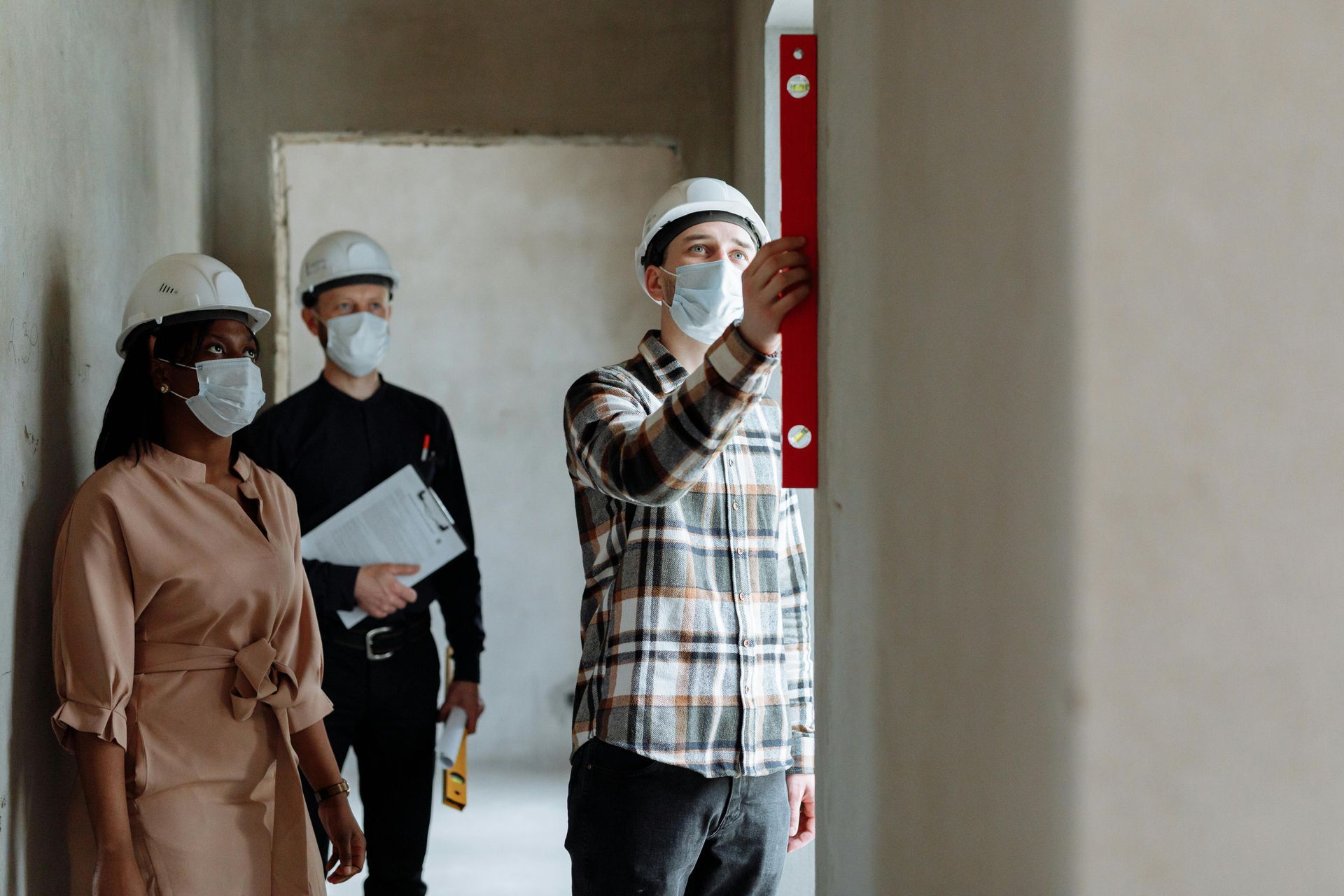 Three people in hard hats and masks inspecting an indoor construction site with a spirit level.