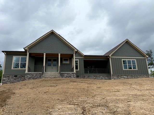 New gray house with stone accents and a front porch on a bare dirt lot under cloudy skies