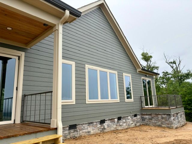 Modern gray house exterior with wood porch, large windows, and stone foundation under cloudy sky