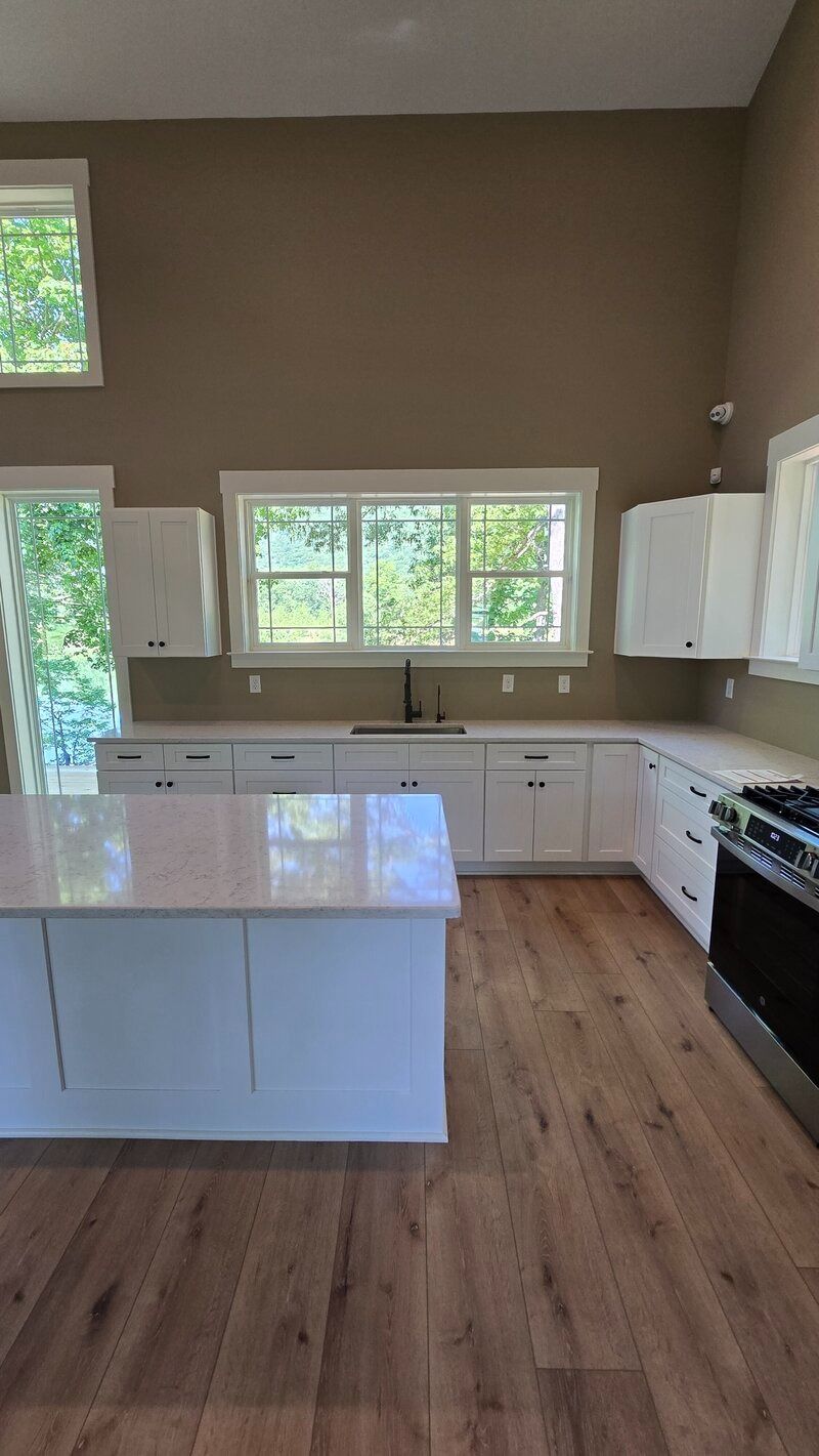 White kitchen with wooden floors. White cabinets, countertops, and island. Brown walls, windows, and stainless steel range.