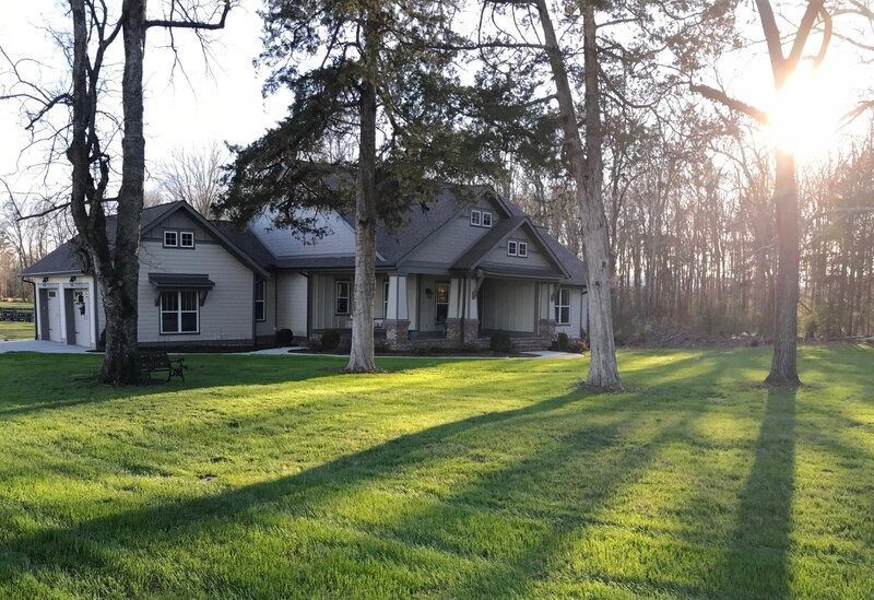House with a gray exterior, front porch, and garage, on a grassy lawn with trees; sunlight in the background.