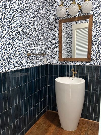 Blue and white leaf patterned wallpapered bathroom with blue vertical tiled walls, wooden floor, and a pedestal sink.