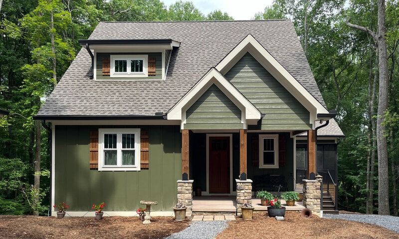 Green-sided house with a dark roof, brown door, and stone and wood accents, surrounded by trees.