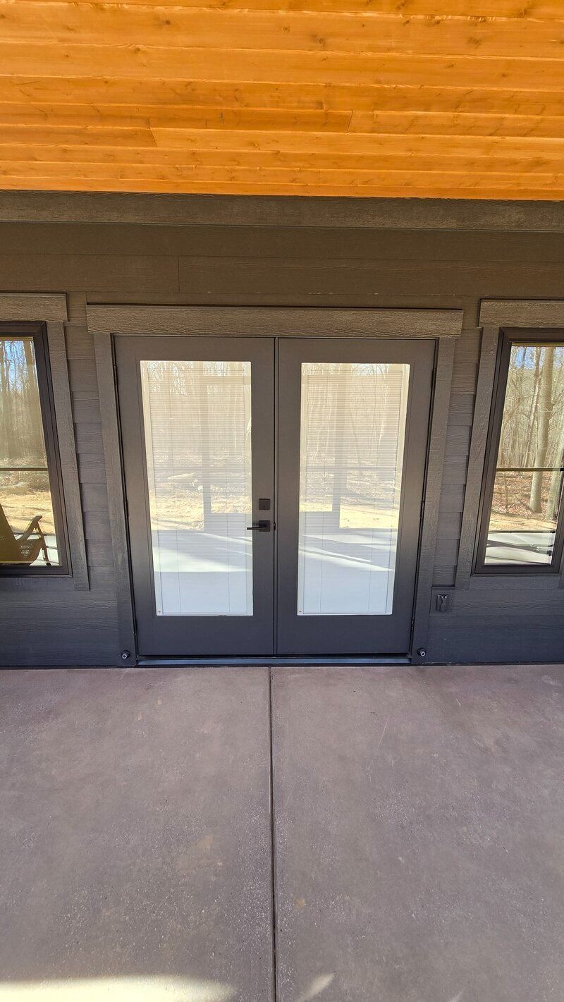French doors on a dark gray building, set in a paved patio with wooden ceiling, and windows on each side.