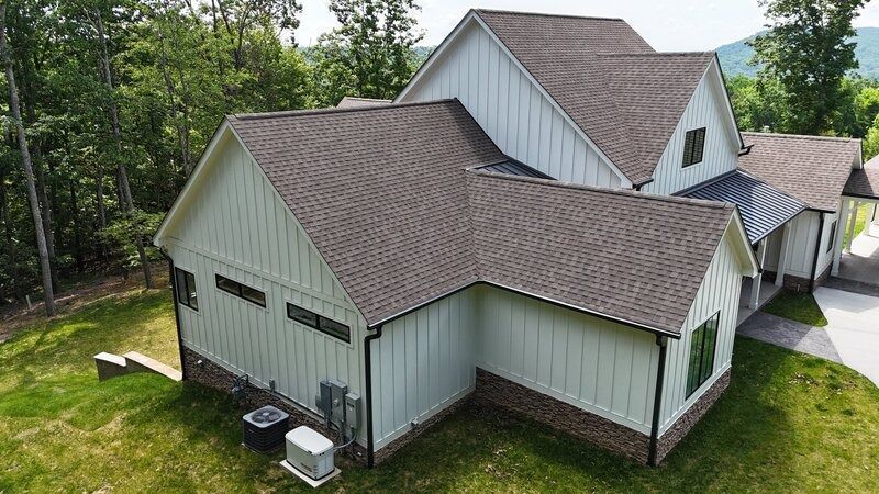 White house with brown roof, green trim, and brick base. Exterior view in a wooded area.