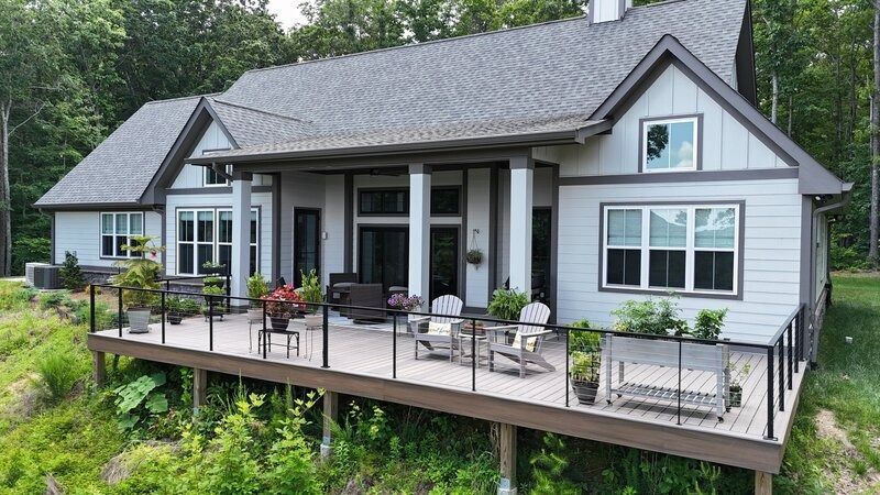 Light gray house with a wooden deck overlooking a grassy area, surrounded by trees.