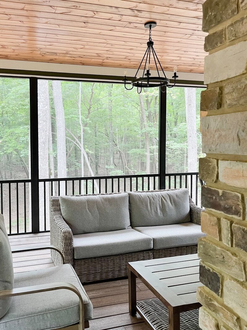 Screened porch with woven sofa, brick wall, and chandelier, overlooking a wooded area.