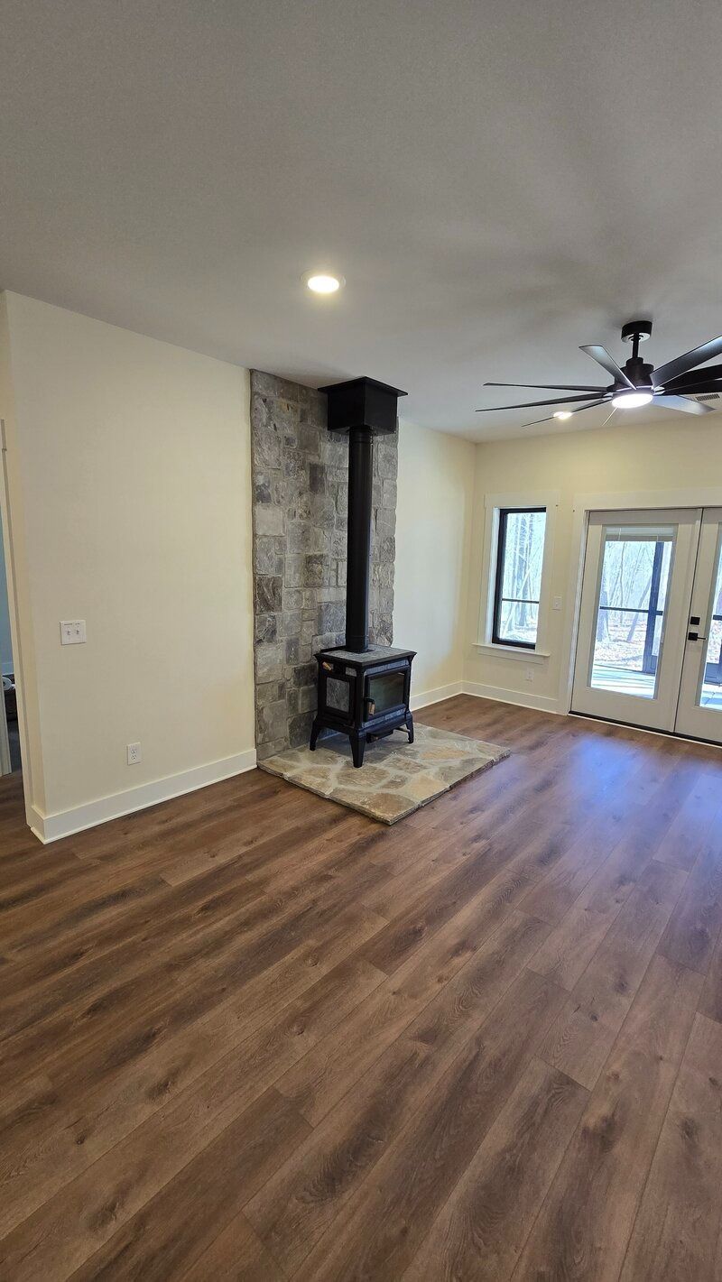 Living room with a wood-burning stove and a stone accent wall, wood-look flooring, and a ceiling fan.