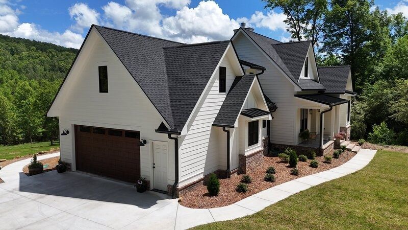 White house with black roof, brown garage door, and landscaping, against a backdrop of trees and a hillside.