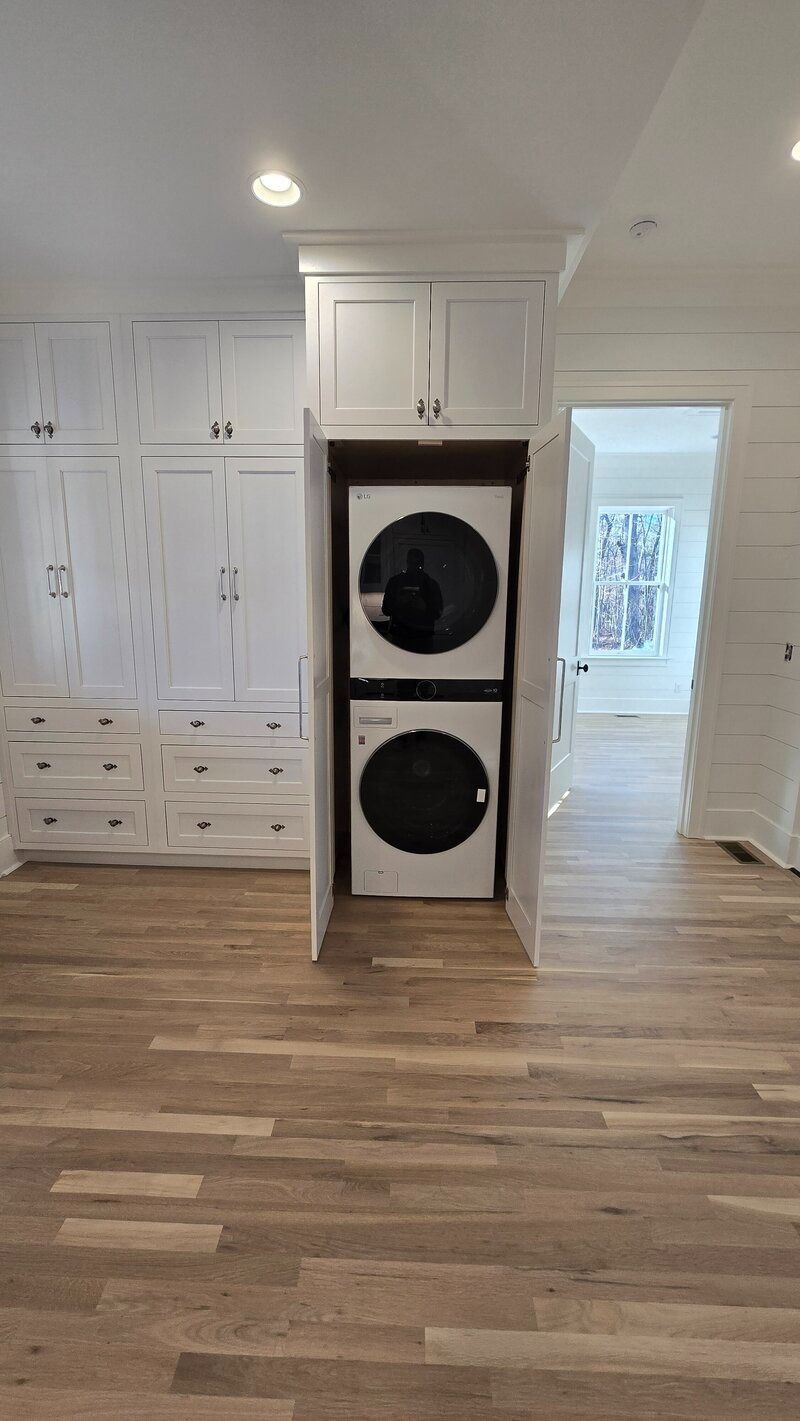 Stackable washer and dryer unit in a white-cabinet laundry room. Wooden floor, doorway to another room.