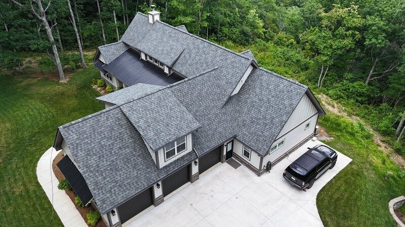 Aerial view of a gray-roofed house with a black car parked in front of the garage. Lush green trees surround the home.