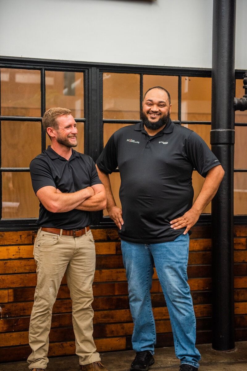 Two men, one with arms crossed, the other with hands on hips, stand in front of windows and a brick wall.