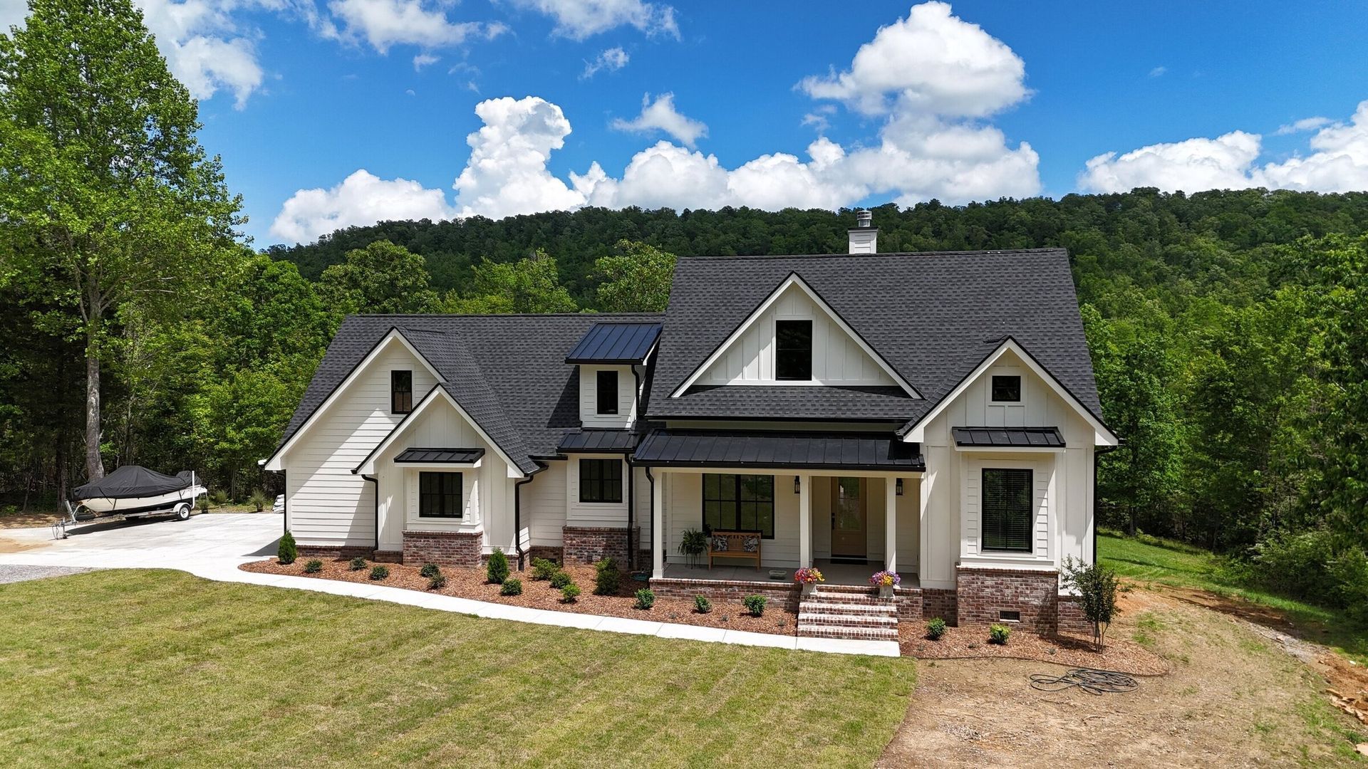 White farmhouse with black roof, brick accents, and a lush green background.