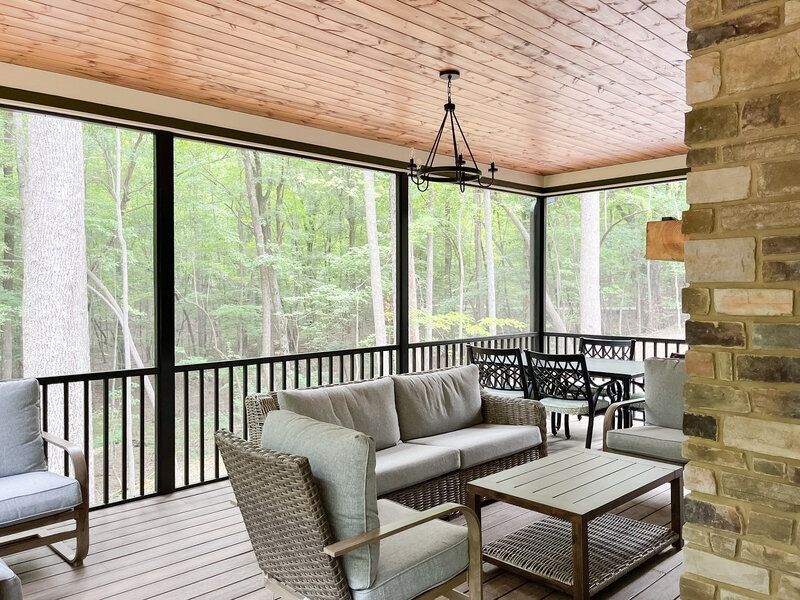 Screened-in porch with outdoor furniture, overlooking a wooded area. The ceiling is wood, the walls are screened.