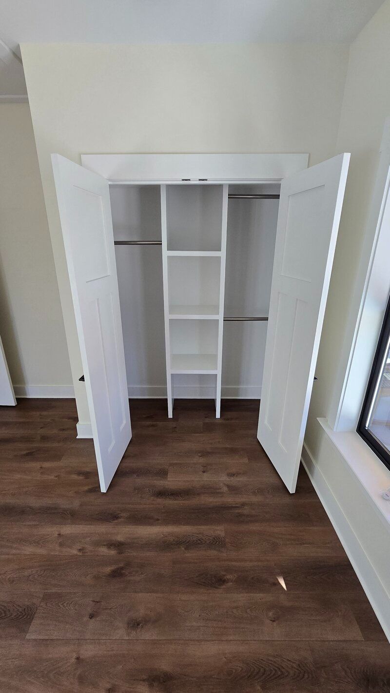 White closet with open doors, shelves, and hanging rods; set in a room with wood flooring.