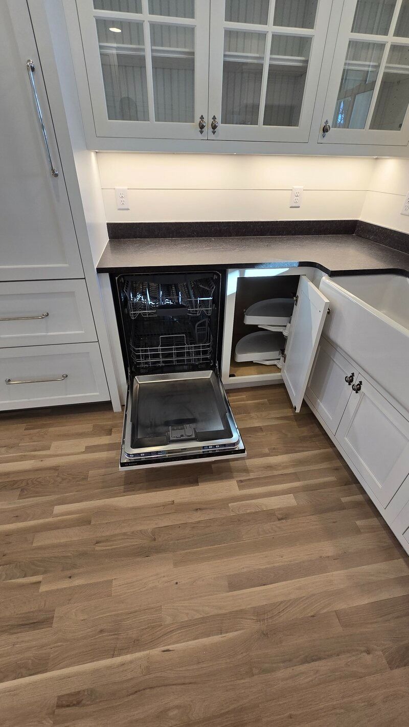 A kitchen with an open dishwasher and cabinetry. Hardwood floors, black countertops, and white cabinets are seen.