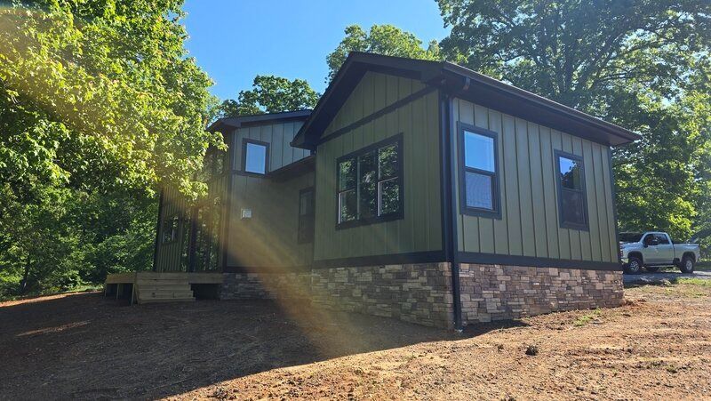Green house with stone base and dark trim, in a wooded setting under a blue sky.