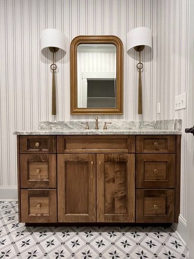 Bathroom vanity with wood cabinets, gold-framed mirror, marble countertop, and patterned tile floor.
