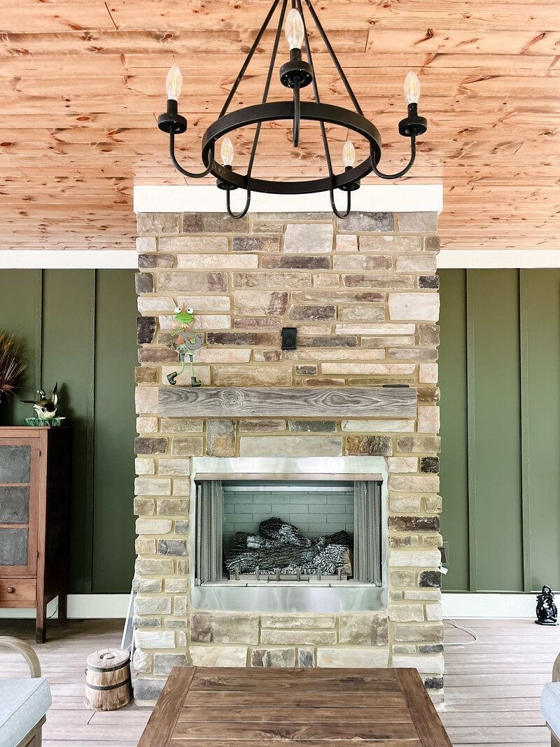 Stone fireplace with a chandelier, on a porch with wood ceiling and green wall.