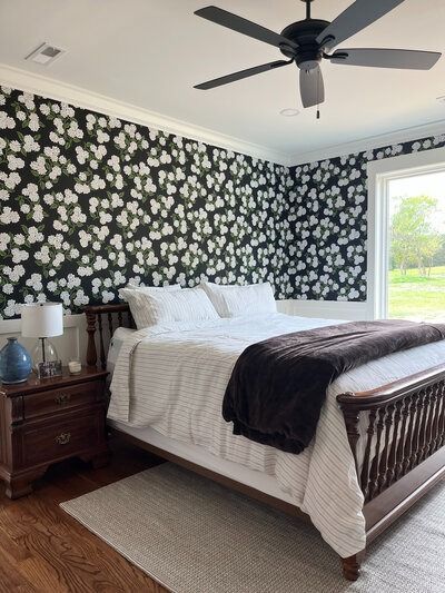 Bedroom with patterned wallpaper, dark bed frame, white bedding, nightstand, ceiling fan, and window.