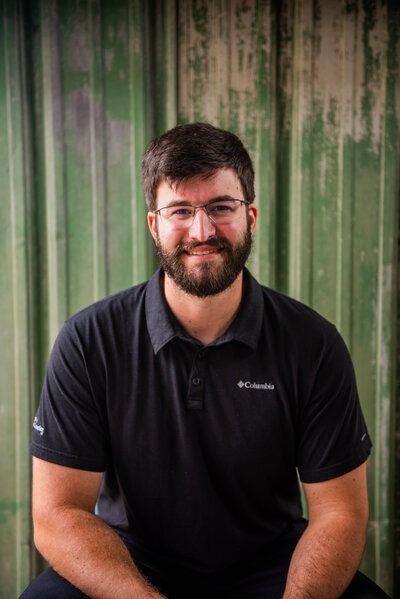 Man with glasses and beard smiles, wearing black Columbia shirt, in front of green textured wall.