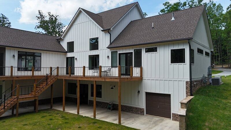 White farmhouse with a brown roof and deck. Includes a garage and is surrounded by trees.