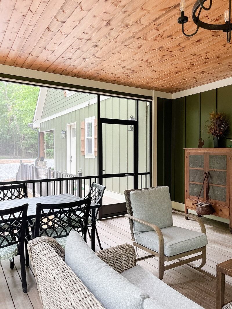 Cozy screened porch with wood ceiling, green walls, and outdoor furniture overlooking a green house.