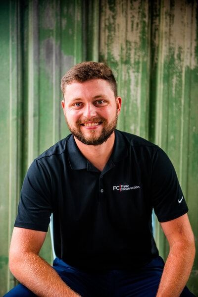 Man with beard in black polo shirt smiles in front of a green wall.