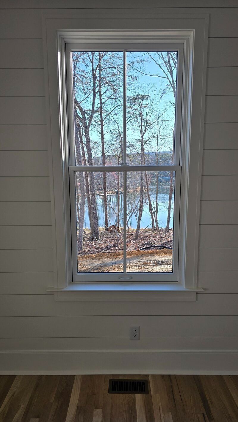 Window looking out onto a lake with trees. White walls, wooden floor, bright outside.