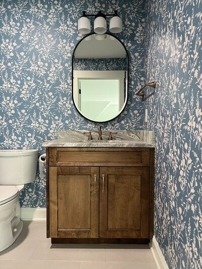 Powder room with blue floral wallpaper, wood vanity, oval mirror, and toilet.