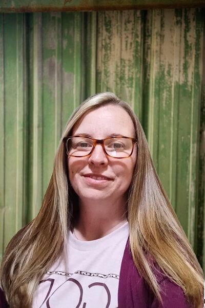 Woman with long blonde hair, glasses, and purple sweater smiling in front of green background.