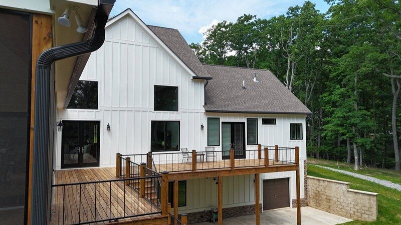 White two-story house with a wooden deck and brown roof, set against a backdrop of trees and a cloudy sky.